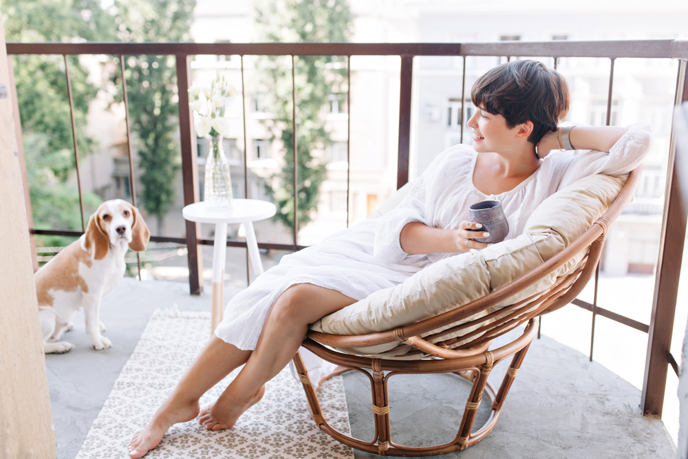 Relaxed barefooted girl in white dress sitting in chair on balcony and holding cup of tea. Adorable brunette woman drinking coffee on terrace and looking with smile at beagle dog.