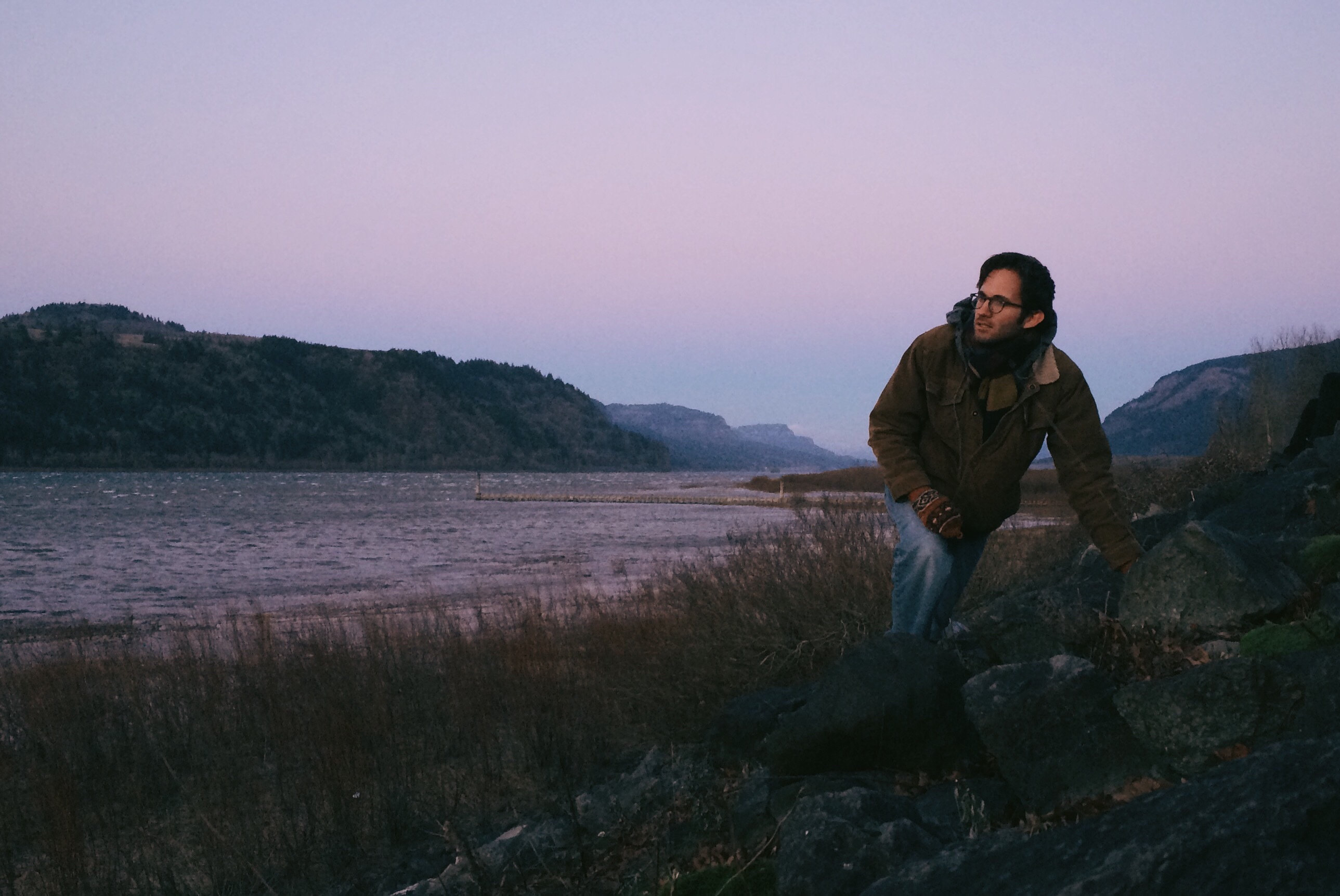 A young man leans on a rock as he stares out into the sunset by the lake at Rooster Rock State Park outside of Portland, Oregon.