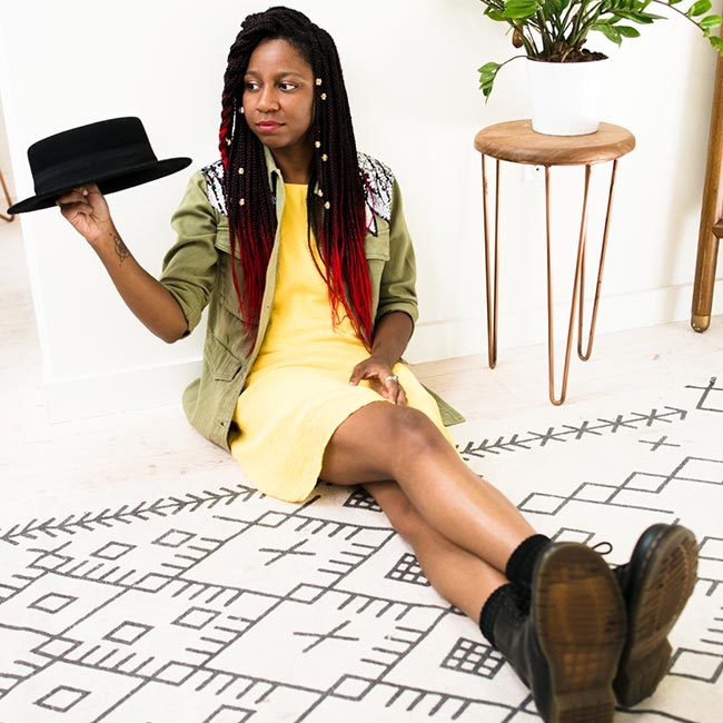 gerilyn hayes sits on wood floor and patterned carpet, and holds black hat in right hand