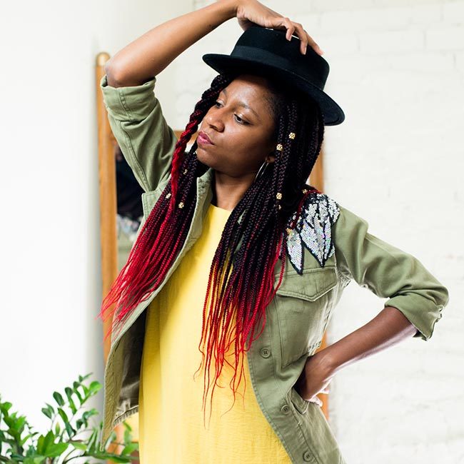 gerilyn hayes wears yellow dress and sequined military-style jacket, and puts black hat onto of ombre red box braids