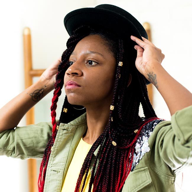 gerilyn hayes wears yellow dress and sequined military-style jacket, and puts black hat onto of ombre red box braids