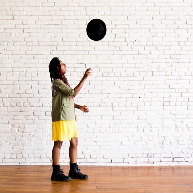 naturallycurly sr copywriter gerilyn hayes gerilyn hayes points at camera while wearing a yellow dress, sequined military-style jacket, black Dr Marten boots, and a black hat onto of ombre red box braids