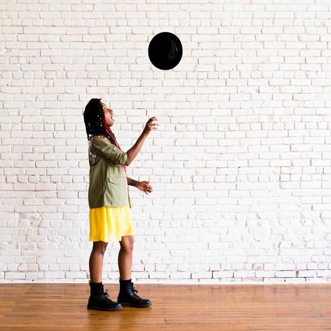 gerilyn hayes points at camera while wearing a yellow dress, sequined military-style jacket, black Dr Marten boots, and a black hat onto of ombre red box braids