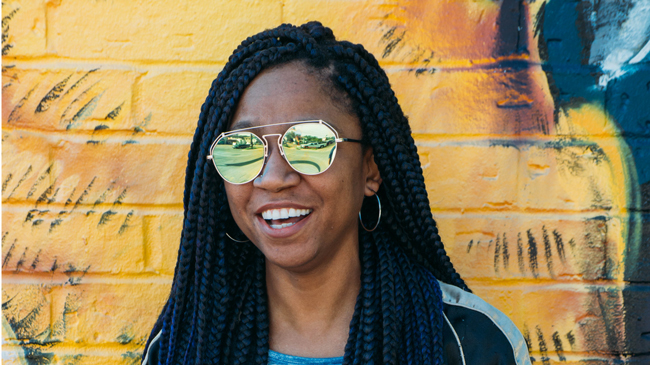 Gerilyn flashes a smile against a bright yellow brick background that constrasts with her indigo braids