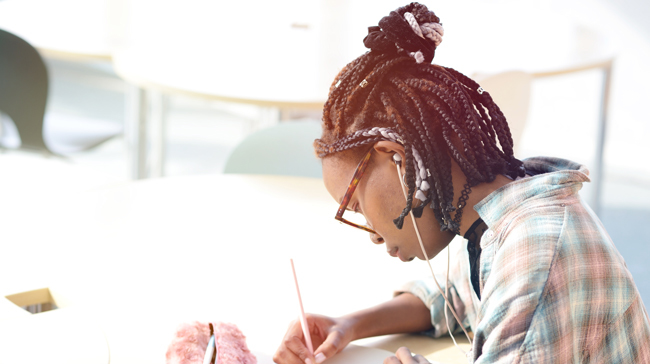 A young black woman with short multicolored box braids takes some notes with a pad and pen